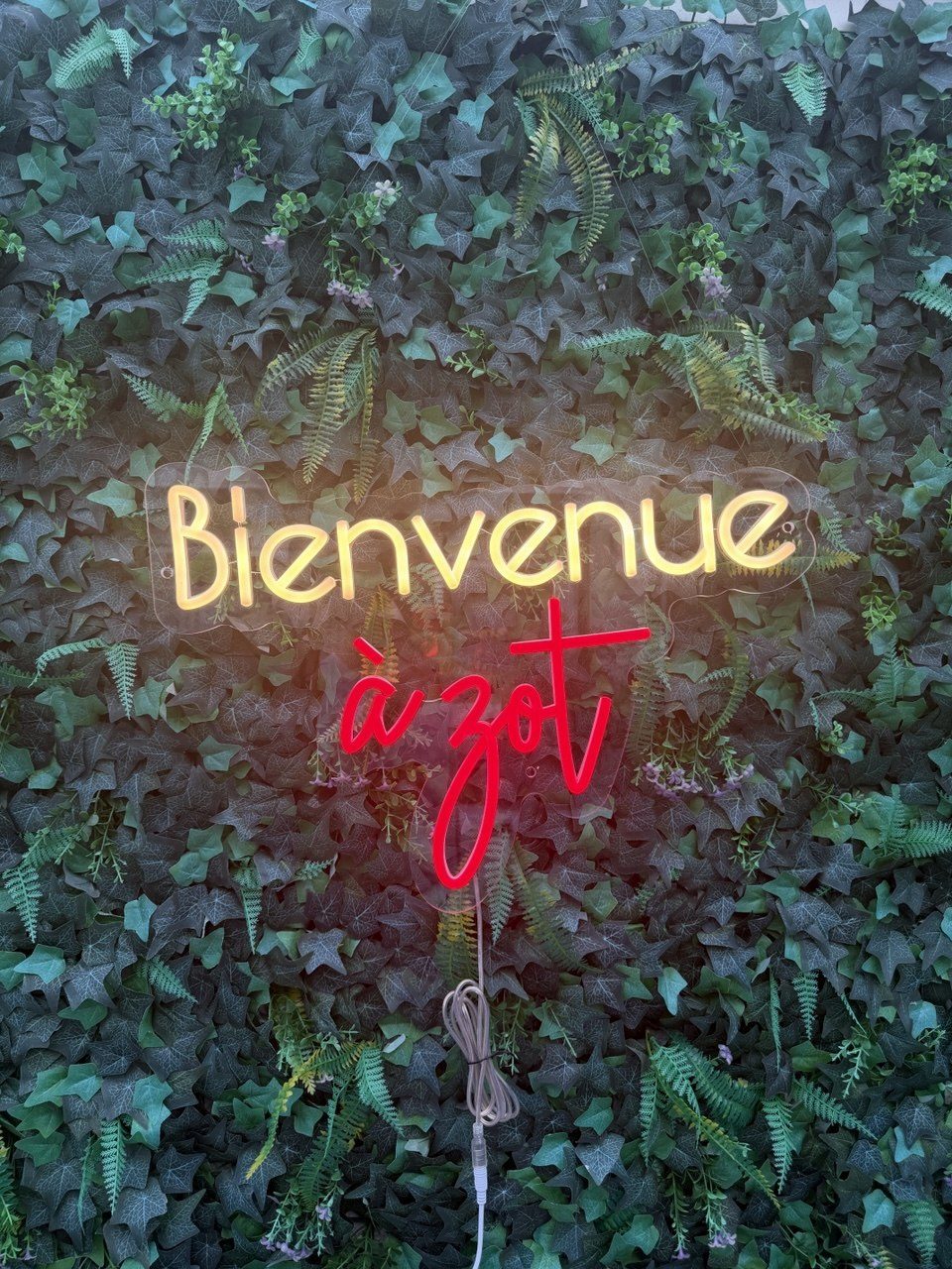 Neonbord met de tekst Bienvenue a zot in warmwit en rood tegen een plantenwand